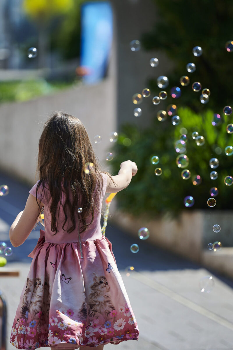 Eventfotografie Düsseldorf Sonnenrad-Festival 2023 - kleines Mädchen mit langen dunklen Haaren im rosafarbenen Kleid pustet Seifenblasen
