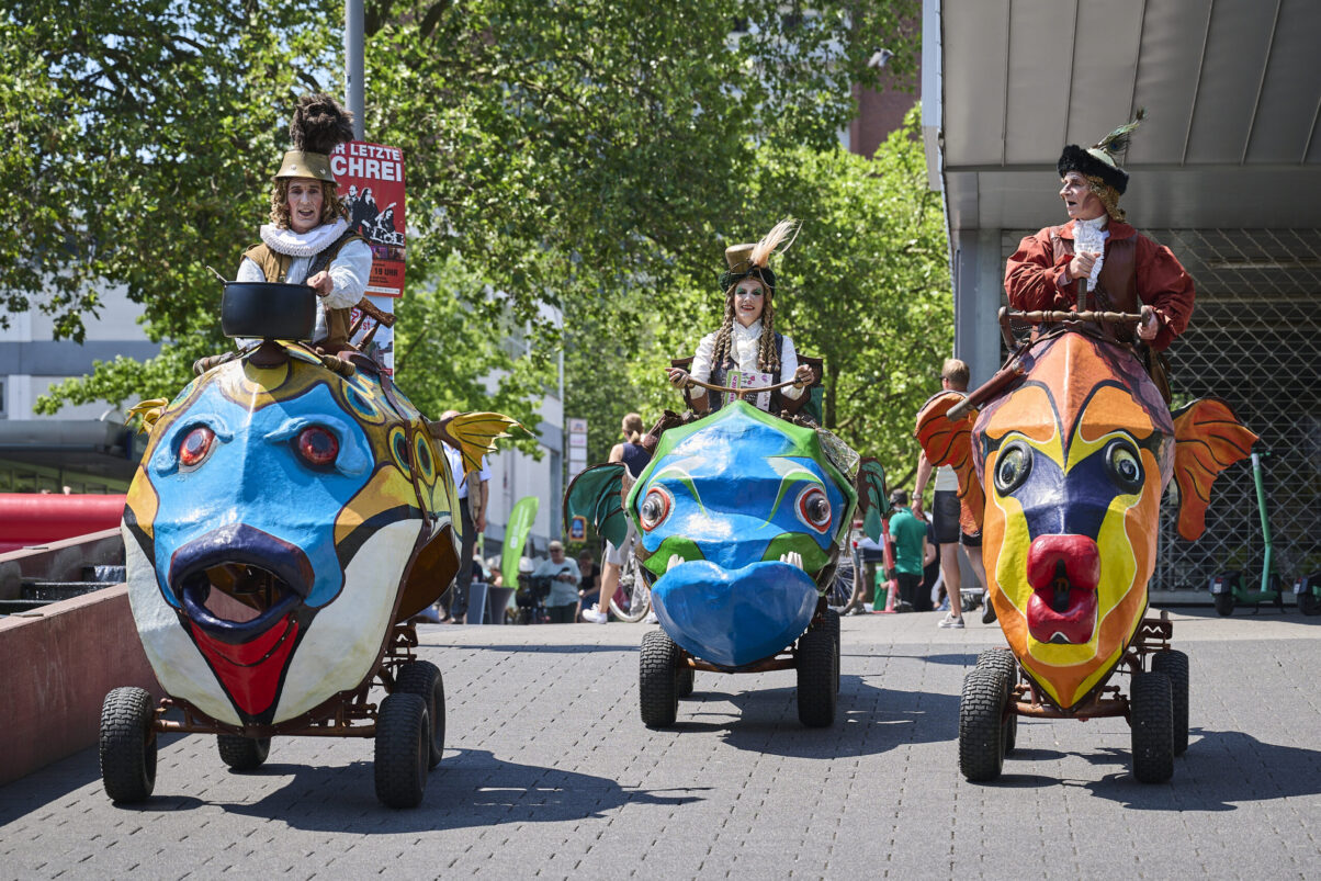 Eventfotografie Düsseldorf Sonnenrad Festival 2023 - altertümlich verkleidete Schausteller in fahrenden großen und bunten Fischen aus Pappmasche