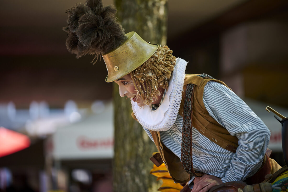Eventfotografie Düsseldorf Sonnenrad Festival 2023 - verkleideter Schausteller mit braunen Federn auf einem Metallhut in die Zuschauermenge schauend