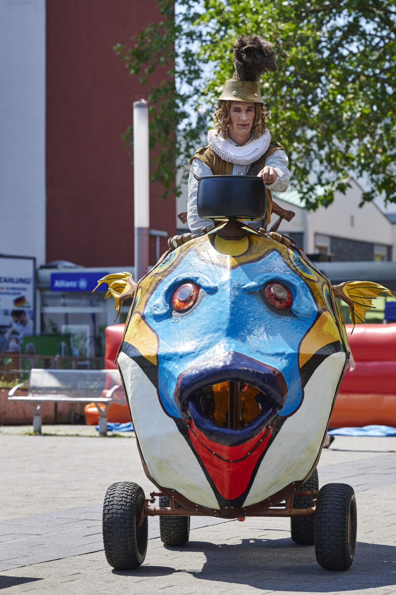 Eventfotografie Düsseldorf Sonnenrad Festival 2023 - Schausteller in barocker Kleidung mit Federhut fährt mit einem übergroßen bunten Fisch aus Pappmasche über den Veranstaltungsplatz