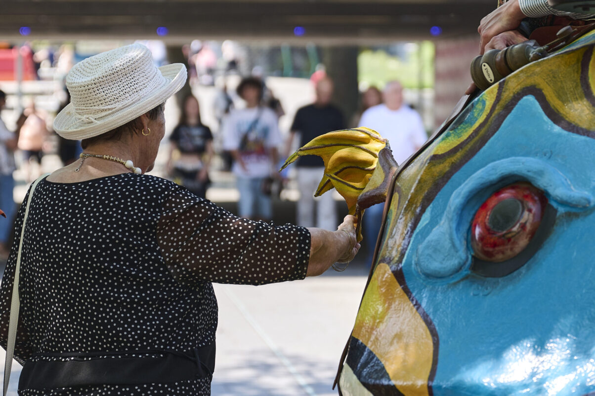 Eventfotografie Düsseldorf Sonnenrad Festival 2023 - ältere Dame mit weißem Hut vor großem bunten Fisch aus Pappmache