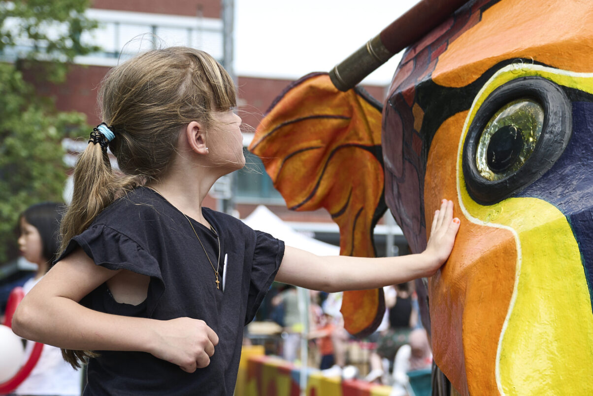 Eventfotografie Düsseldorf Sonnenrad Festival 2023 - dunkelhaariges Mädchen vor großem bunten Fisch aus Pappmache