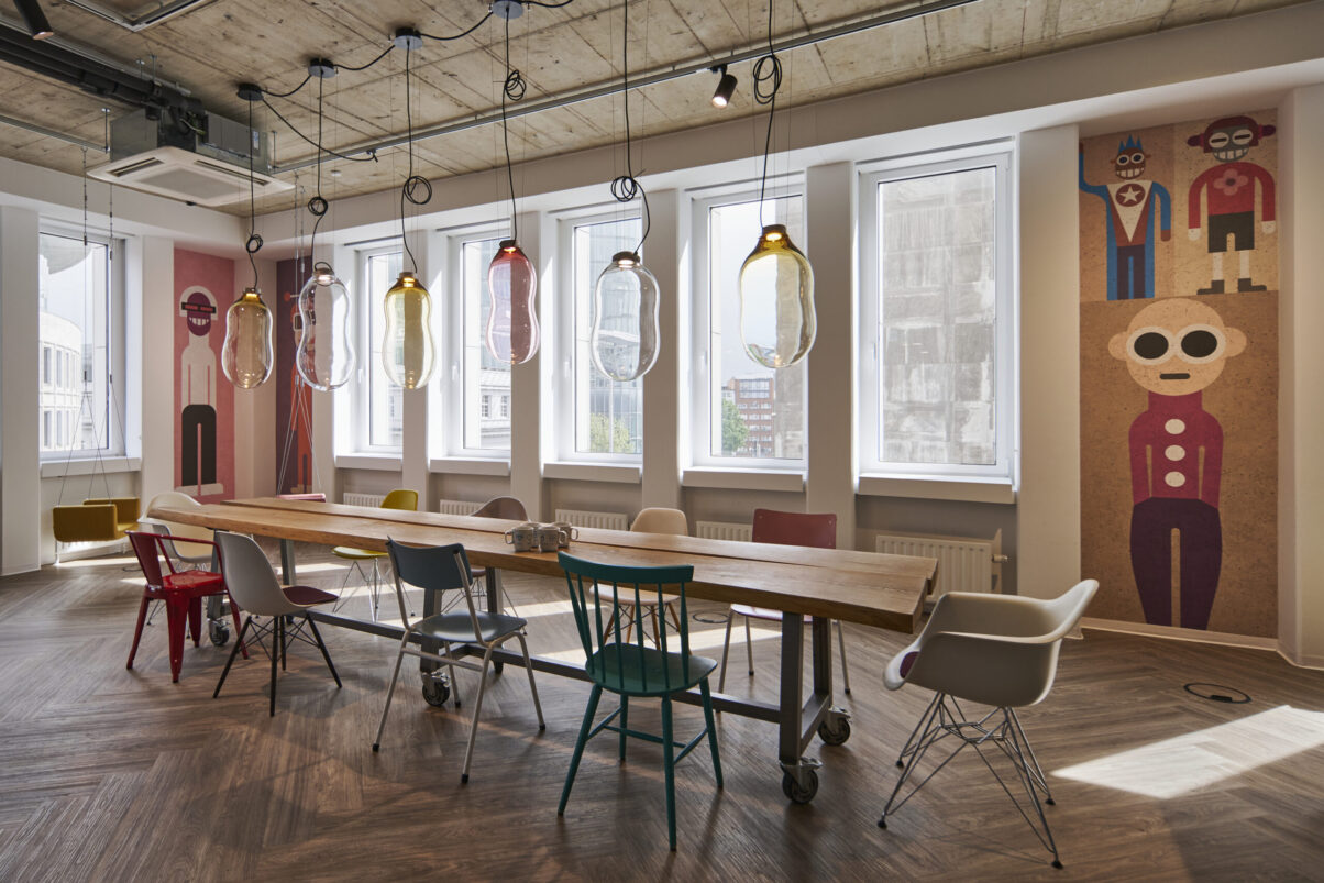 Architektur- und Interieurfotografie Düsseldorf Auxmoney, stilvoller Mitarbeitertreffpunkt, langer außergewöhnlichem Holztisch mit vielen verschiedenen bunten Stühlen, sowie bunten Glaslampen und Wandmotiven