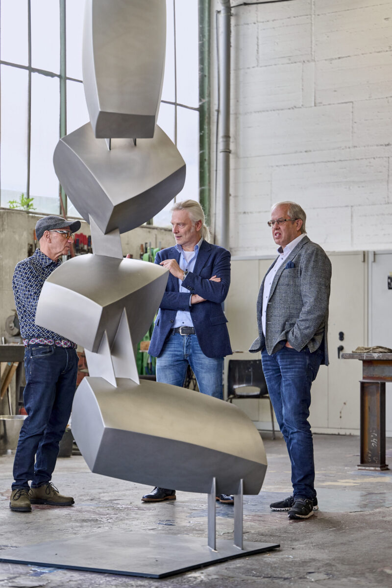 Studio Talk mit Thomas Schönauer - der Künstler im zugewandten Gespräch mit zwei Herren - Kunstobjekt aus Metall im Vordergrund