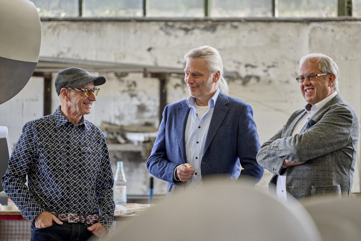 Studio Talk mit Thomas Schönauer - der Künstler im zugewandten Gespräch mit zwei Herren - Kunstobjekt aus Metall im Hintergrund