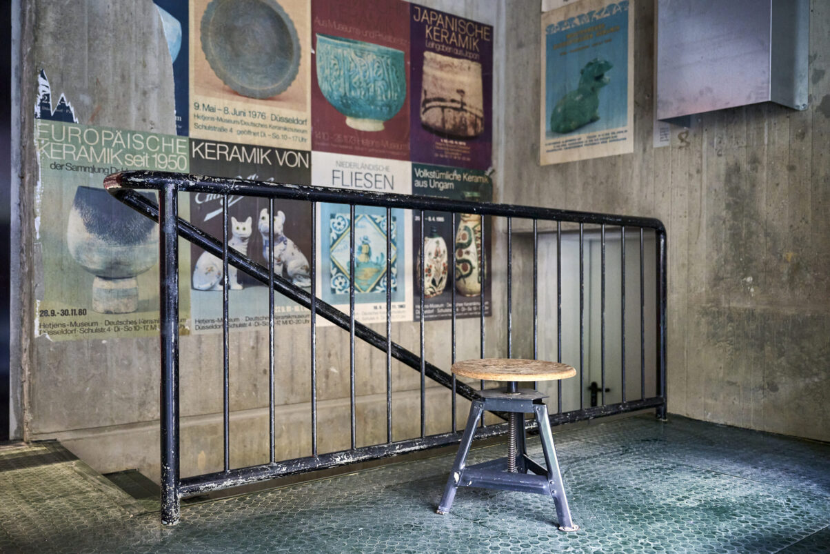 Architektur- und Interieurfotografie Düsseldorf Kulturhaus Süd, lichtdurchflutete Töpferwerkstatt, Stillleben mit Drehschemel und Treppengeländer, alte Keramikposter an der Wand im Hintergrund