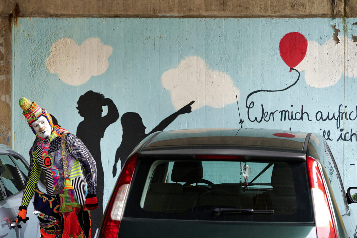 Pop up Artistik Festival, farbenprächtig gekleideter Pantomime Clown, zwischen Autos vor einer bunten Graffitiwand