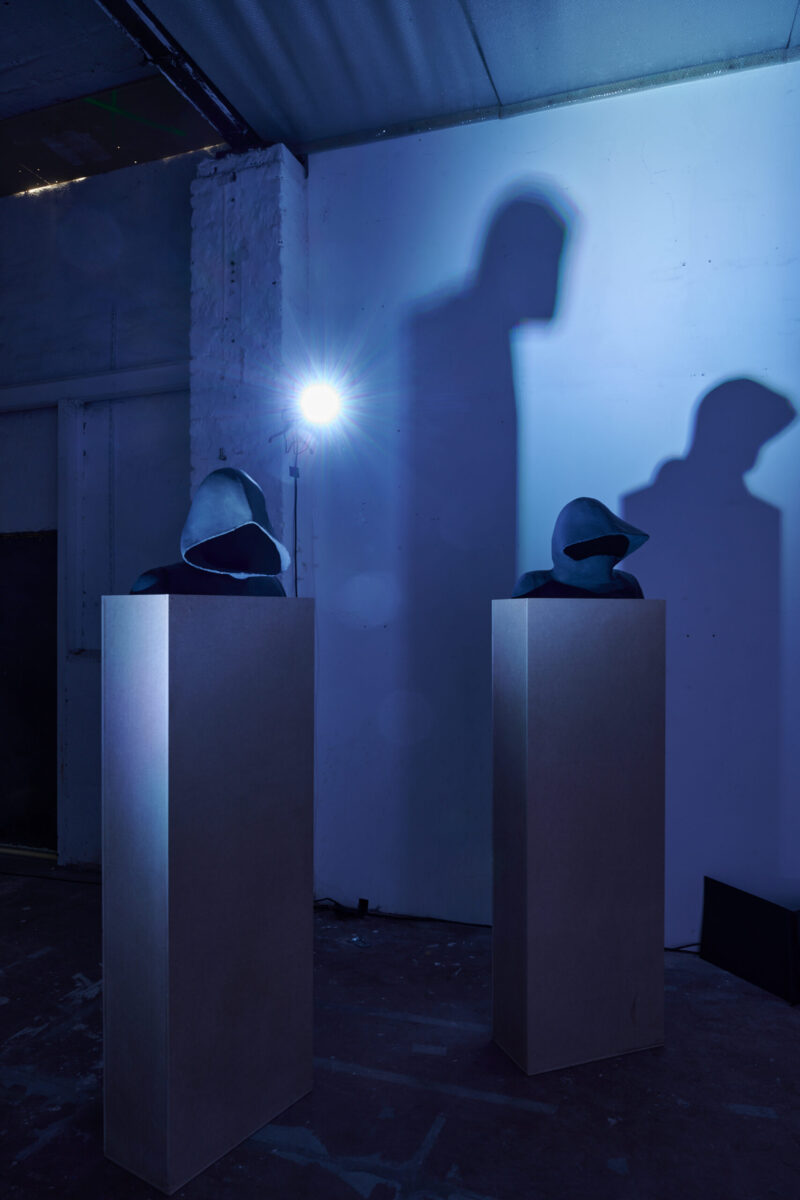 Eventfotografie Düsseldorf Ausstellung "72 Stunden - ein Zeitraum" von Corinna Bernshaus - zwei Büsten mit Kapuze, blaue Tonskulpturen auf weißen Säulen und Ihr Schattenspiel auf Wand