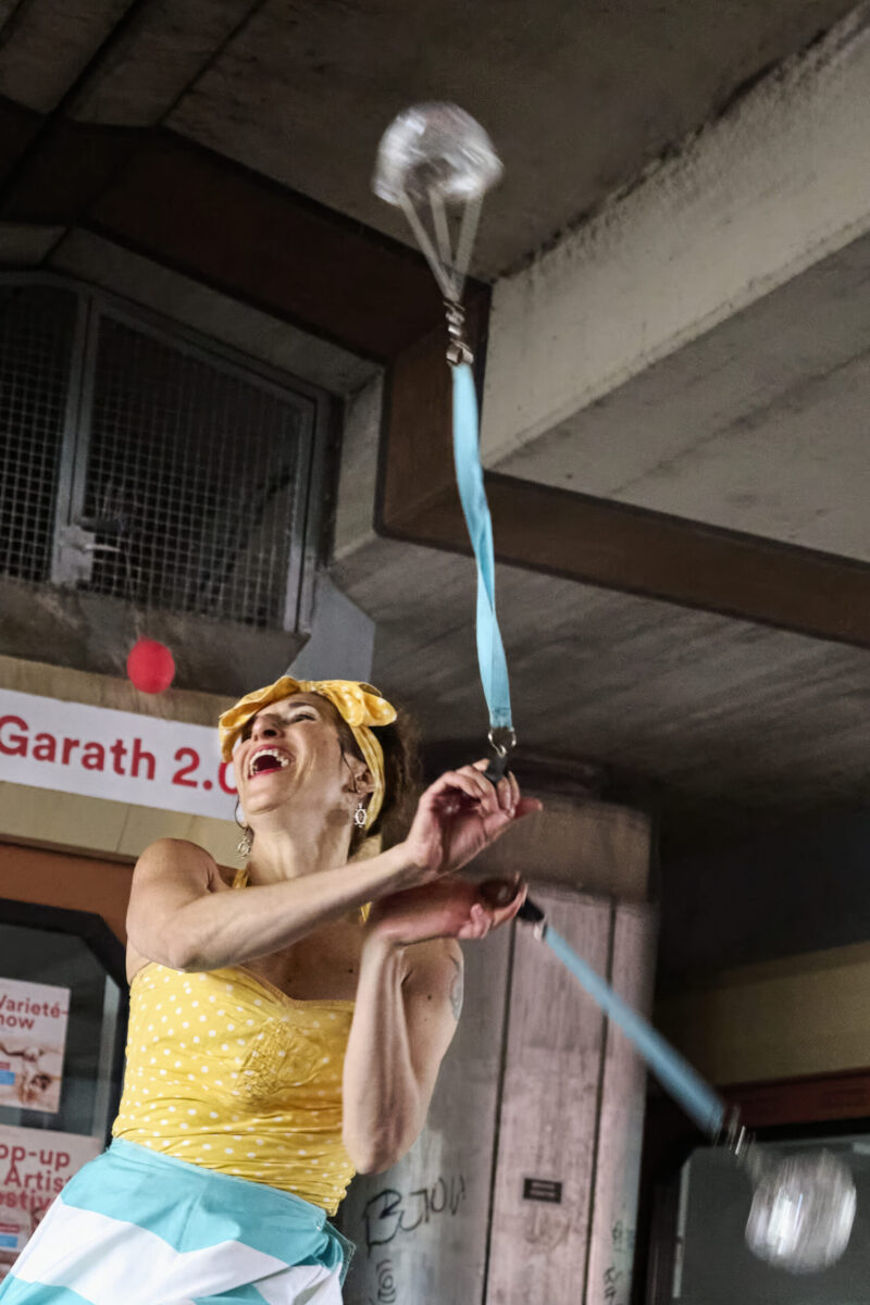 Pop up Artistik Festival, Nahaufnahme einer Artistin, lachend, zwei mit Wasser gefüllte Glasgefäße an Bändern jonglierend