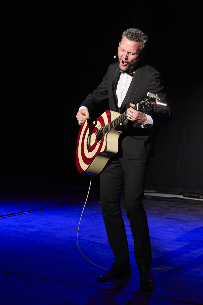 Eröffnungsgala Kulturhaus Süd - Comedian im schwarzen Scmoking spielt auf einer elektrischen Gitarre. Schwarzblaues Bühnenbild.