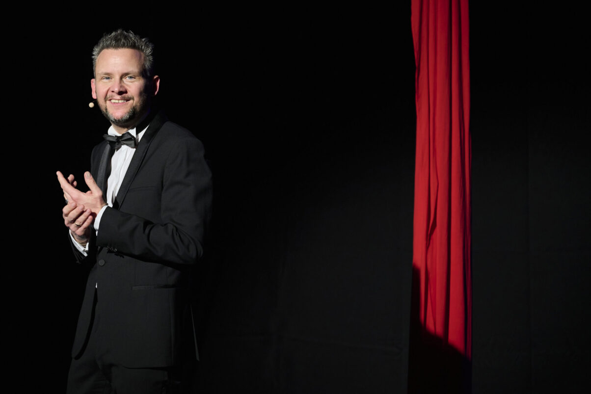 Eröffnungsgala Kulturhaus Süd - Comedian im schwarzen Smoking mit viel Humor schaut ins Publikum. Rotes Tuch hängend im Hintergund. Schwarzes Bühnenbild.