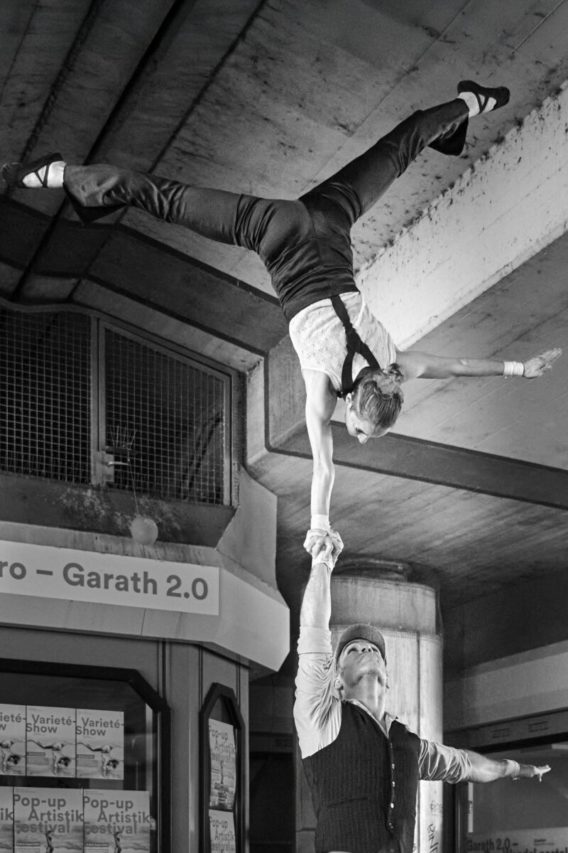 Pop up Artistik Festival, Artistenpaar, er stemmt sie im Handstand auf einer Hand in die Höhe, Foto in sw
