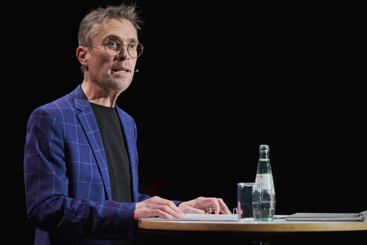 Eröffnungsgala Kulturhaus Süd - Comedian mit Brille und kariertem Sakko steht am Rednerpult und schaut ins Publikum