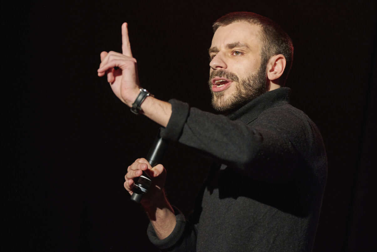 Eröffnungsgala Kulturhaus Süd - Comedian mit Bart im schwarzen Pullover spricht ins Mikrofon und schaut ins Publikum