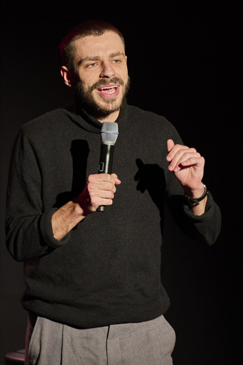 Eröffnungsgala Kulturhaus Süd - Comedian mit Bart im schwarzen Pullover spricht ins Mikrofon und schaut ins nach vorne