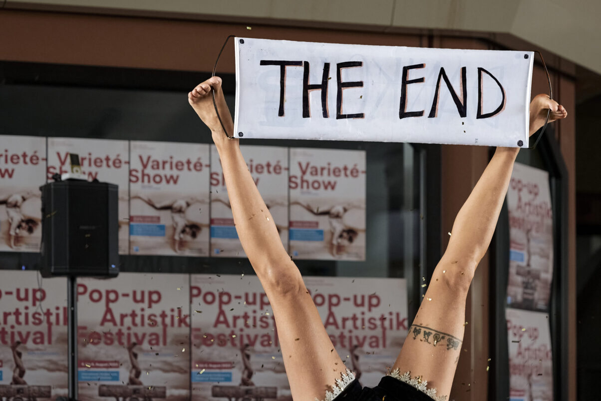 Pop up Artistik Festival, Artistin präsentiert kopfüber ein Schild "Ende" zwischen ihren Füßen, das Ende ihrer Vorstellung
