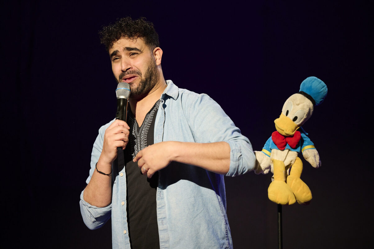 Eröffnungsgala Kulturhaus Süd - Comedian im hellblauen Jeanshemd spricht ins Mikrofon mit Donald Duck Stoffpuppe im Hintergrund