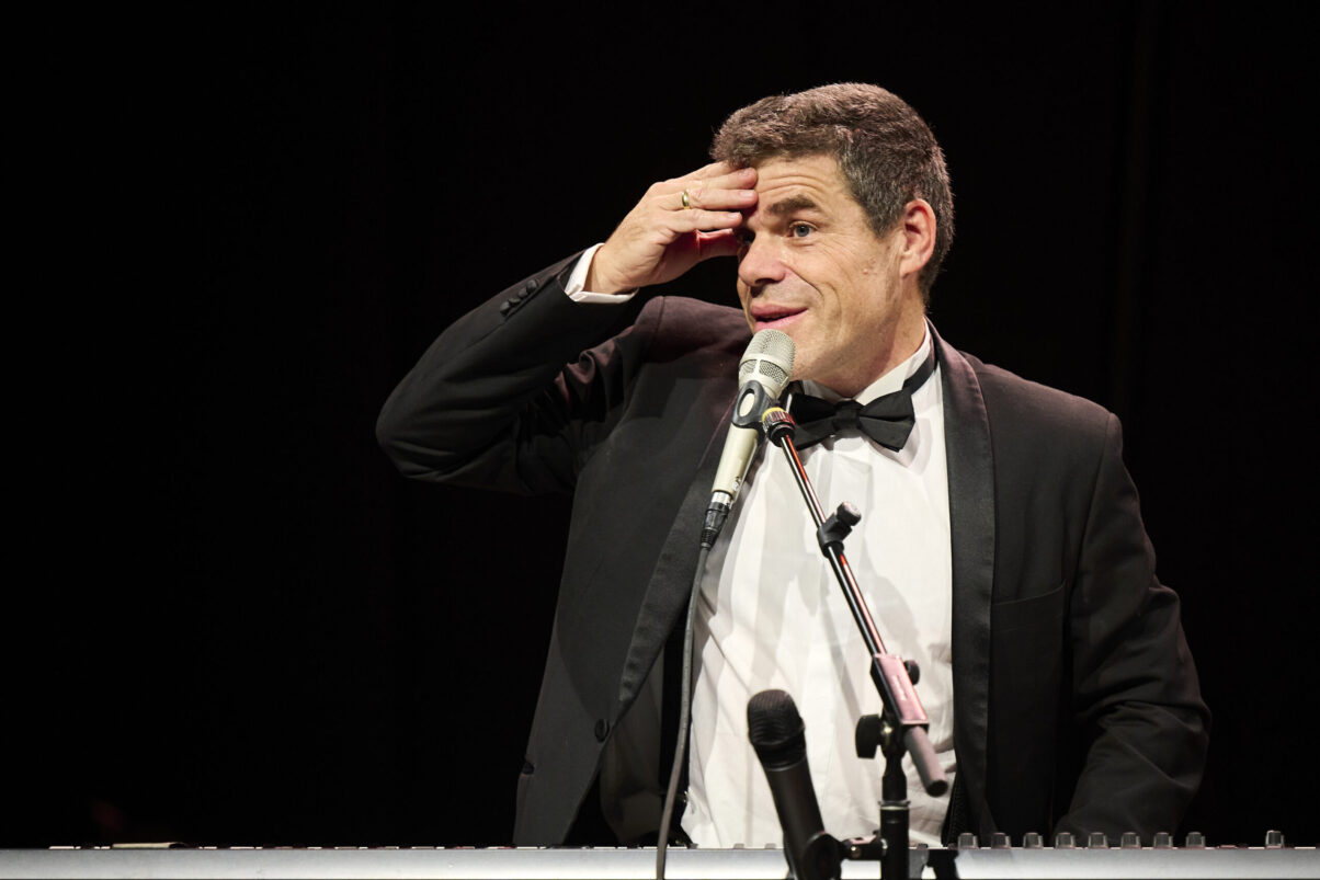 Eröffnungsgala Kulturhaus Süd - Comedian im schwarzen Smoking auf der Bühne am Keyboard schaut ins Publikum