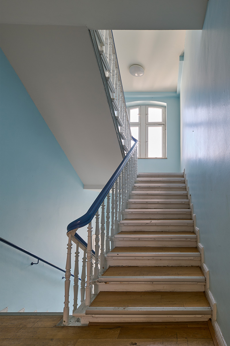 Architektur- und Interieurfotografie Düsseldorf SKFM Sanierung Altbau, Treppenhaus mit hellblauen Wänden und einer alten Treppe mit Parkettstufen und weiß lackiertem Holzgeländer, Handlauf in sattem Blau